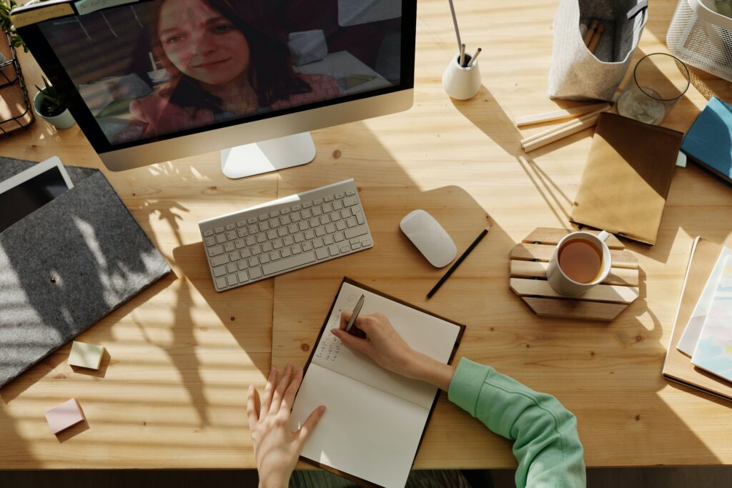 A person sitting at a wooden desk, writing in a notebook, with a cup of coffee to the side. On the computer screen, a virtual meeting is taking place with a woman's face visible, suggesting a remote work session or virtual collaboration.